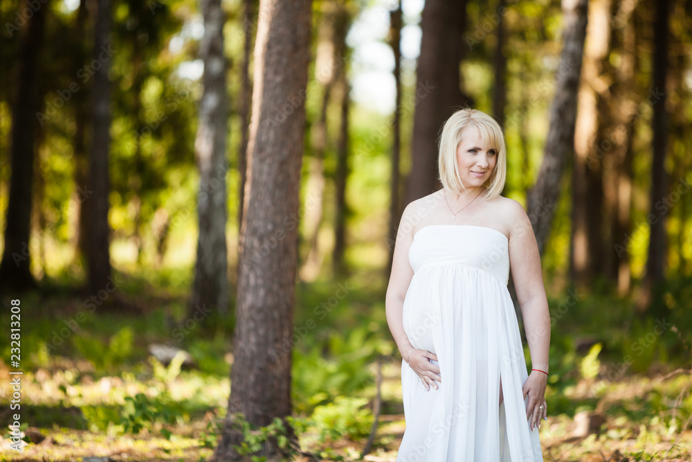 Portrait of beautiful pregnant woman in sheer long white maternity dress looking dreamy in the middle of forest Portrait of beautiful pregnant woman in sheer long white maternity dress looking dreamy in the middle of forest