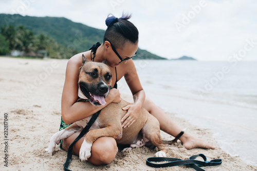 Cheerful asian young woman in eyeglases sitting and hugging her dog on the beach photo
