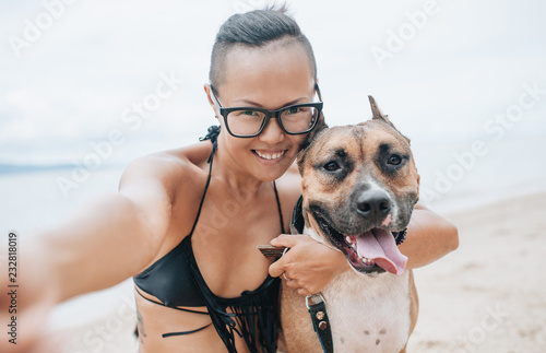 Cheerful asian young woman in eyeglases sitting and doing sekfie with her dog on the beach