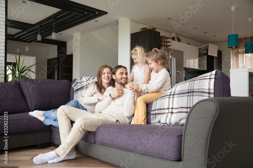 Young attractive married couple with little preschool children sitting on couch in living room at new modern wealthy house. Happy playful parents and joyful kids spending free time or weekend together