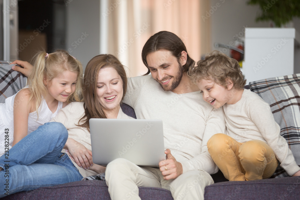 Married couple and little children sitting on couch in living room. Parents with little daughter and son looking at computer and smiling. Happy family together at home, enjoying weekend and free time