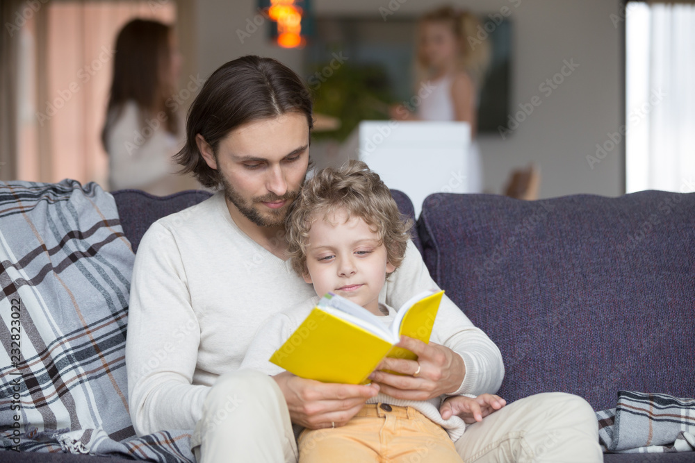 Family with children at home. Little son sitting together with father on couch in living room, mother and daughter out of focus on background. Male read fairytale story book, kid listen with interest Family with children at home. Little son sitting together with father on couch in living room, mother and daughter out of focus on background. Male read fairytale story book, kid listen with interest