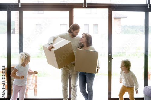 Married young couple enter living room with little children. Family moving at new house. Smiling parents holding unopened cardboard boxes, joyful toddler son and daughter running and playing around