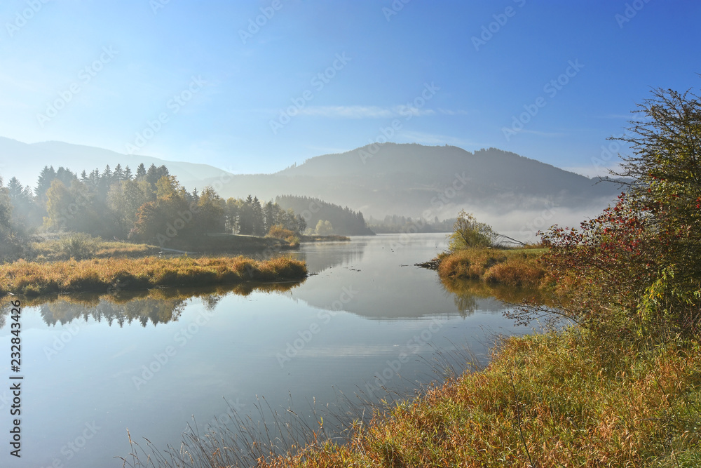 Idyllic morning mood in autumn at the lake Gruentensee in the Allgaeu region (Bavaria, Germany). Lakeshore with colorful grass, bushes and trees. Last fog patches, hills in the background and blue sky