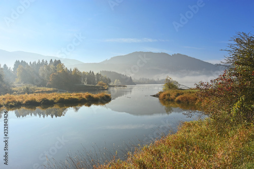 Idyllic morning mood in autumn at the lake Gruentensee in the Allgaeu region  Bavaria  Germany . Lakeshore with colorful grass  bushes and trees. Last fog patches  hills in the background and blue sky