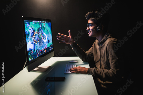 Gamer man pointing with hand on monitor it is challenging the spectator to play. Serious expression, wearing a gamer headset, holding a wired game control at home.