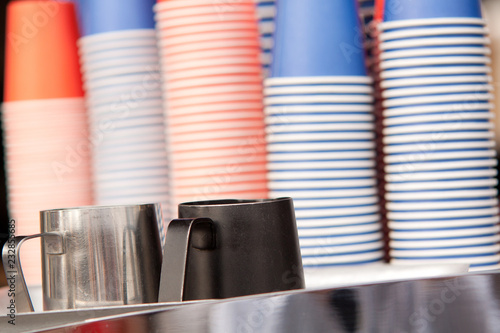 two metal milk jugs and a stack of colored paper cups for making take-away coffee in a cafe photo