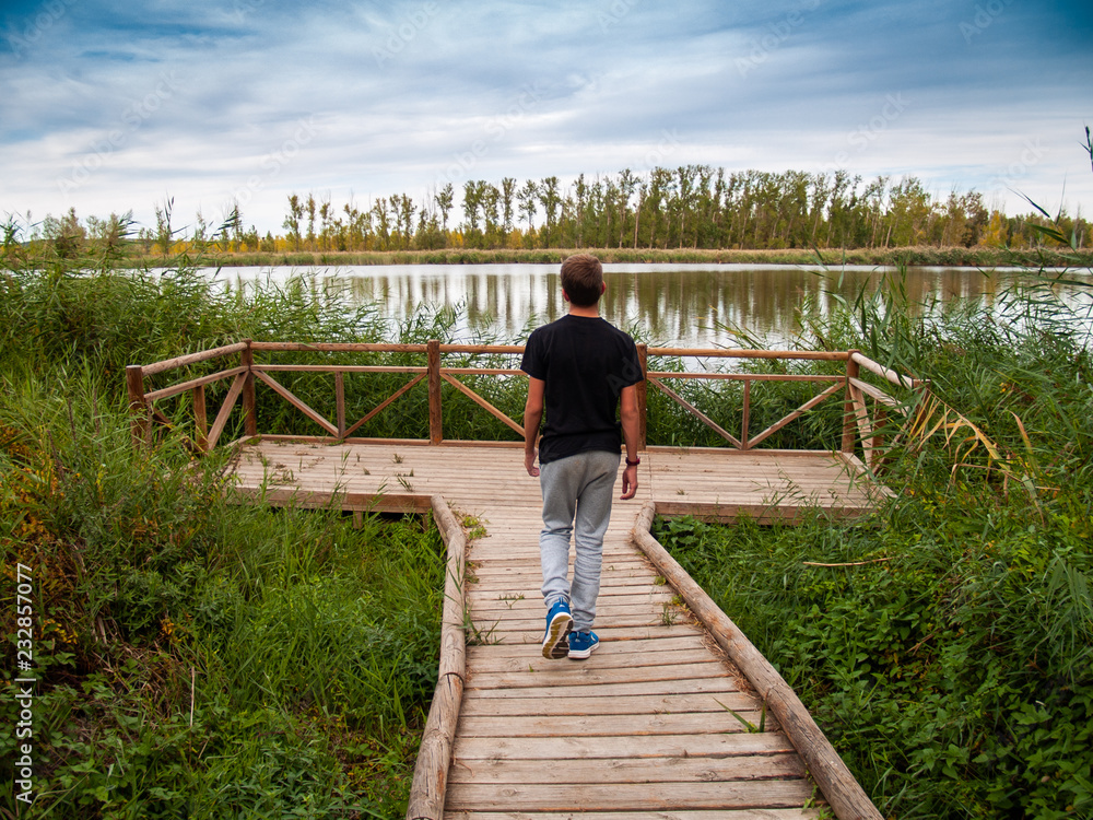 A teenage boy in a viewpoint with a wooden railing contemplating a river in autumn