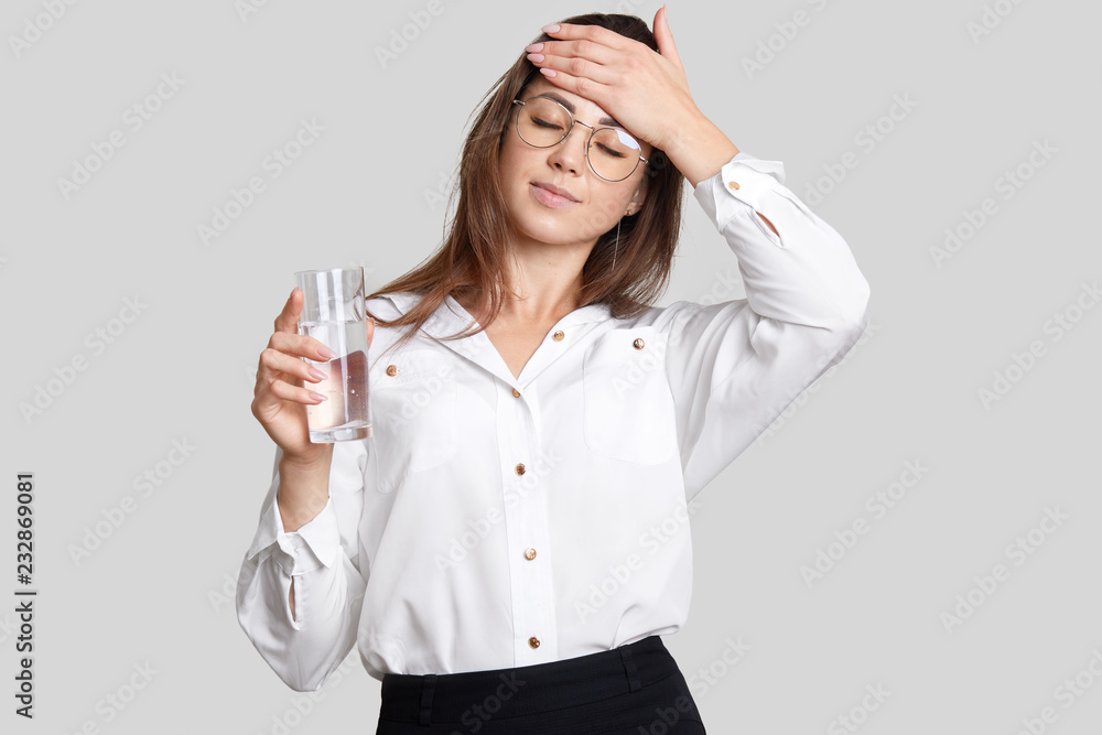 Tired overworked woman keeps hand on forehead, wears transparent glasses, black and white clothes, holds glasses of water, feels thirsty and fatigue, isolated over white background. Weariness Tired overworked woman keeps hand on forehead, wears transparent glasses, black and white clothes, holds glasses of water, feels thirsty and fatigue, isolated over white background. Weariness