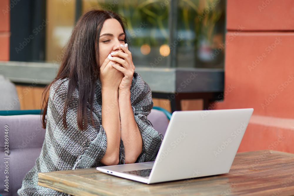 Tired sleepy female entrepreneur yawns in front of modern laptop computer, waist for feedback, works on business report, sits at cozy coffee shop has warm plaid on shoulders. Tiredness and job concept Tired sleepy female entrepreneur yawns in front of modern laptop computer, waist for feedback, works on business report, sits at cozy coffee shop has warm plaid on shoulders. Tiredness and job concept