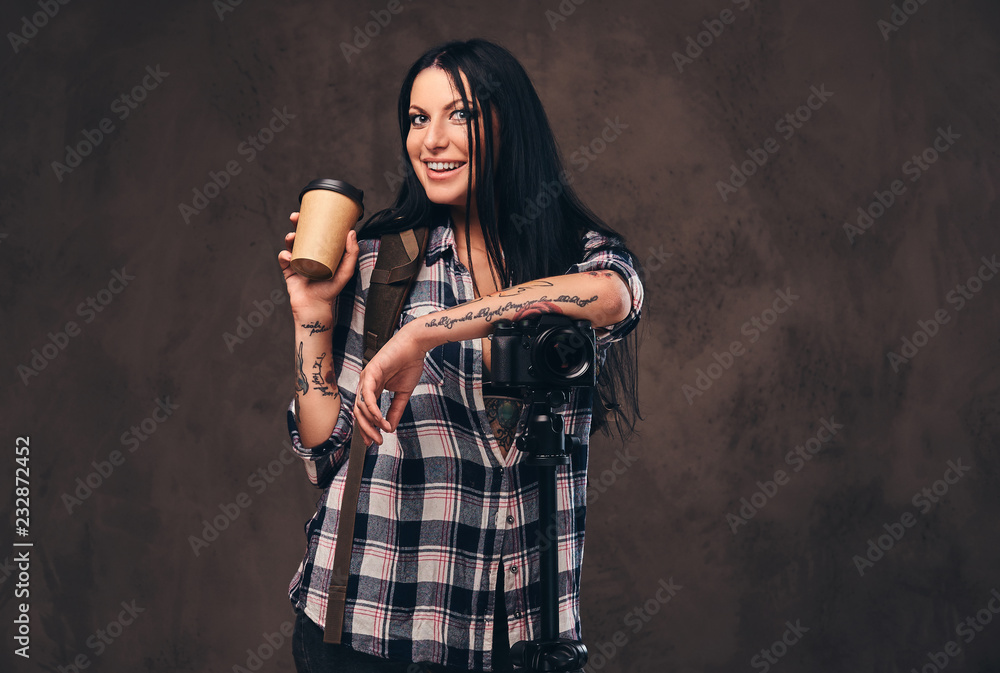 Tattooed girl holding a takeaway coffee while leaning on a camera in a studio.
