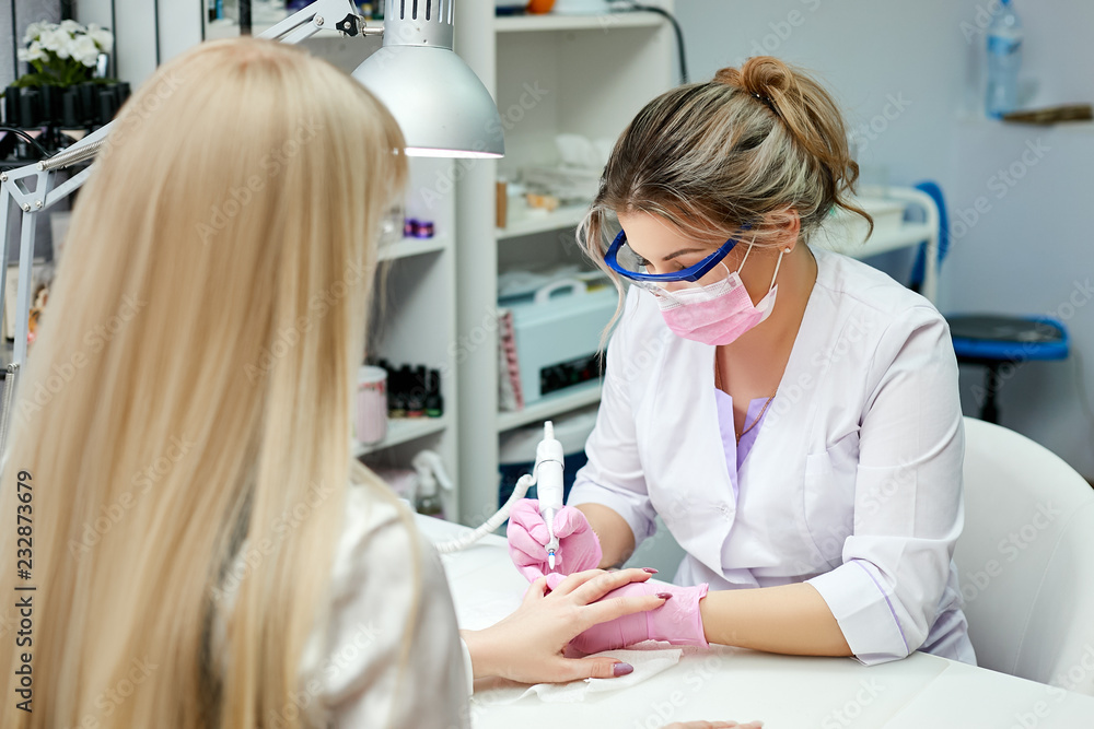 The nail master in lab coat does hardware manicure in the beauty salon. Removal of the gel varnish with a milling cutter. The nail master in lab coat does hardware manicure in the beauty salon. Removal of the gel varnish with a milling cutter.