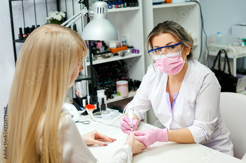 Hardware manicure in the beauty salon. The manicurist in goggles and a pink mask applies a nail drill for manicure on female fingers.