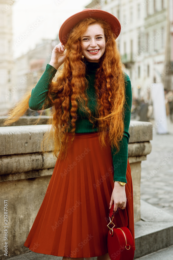 Portrait of young beautiful fashionable happy smiling redhead woman with freckles, very long curly hair, wearing green turtleneck, orange hat, pleated skirt, holding red round bag, posing in street Portrait of young beautiful fashionable happy smiling redhead woman with freckles, very long curly hair, wearing green turtleneck, orange hat, pleated skirt, holding red round bag, posing in street