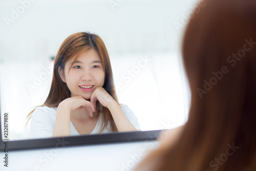 Beautiful of portrait asian young woman examining with face and smile looking on mirror at bedroom, girl beauty of makeup and dressed up with reflection, lifestyle and wellness concept.