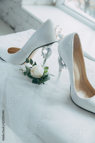 A pair of white wedding shoes with rings on a stool.