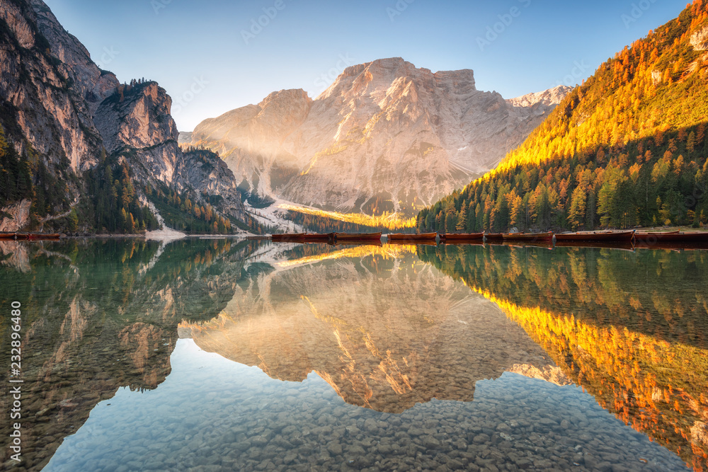 Beautiful Braies lake at sunrise in autumn in Dolomites, Italy. Landscape with mountains, gold sunlight, water with reflection, trees with orange leaves in fall. Travel in italian alps. Dolomiti