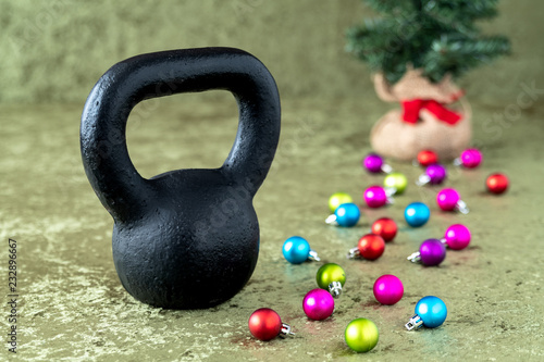 Black kettlebell on a green velvet background with colorful ball ornaments, holiday fitness, Christmas tree in background