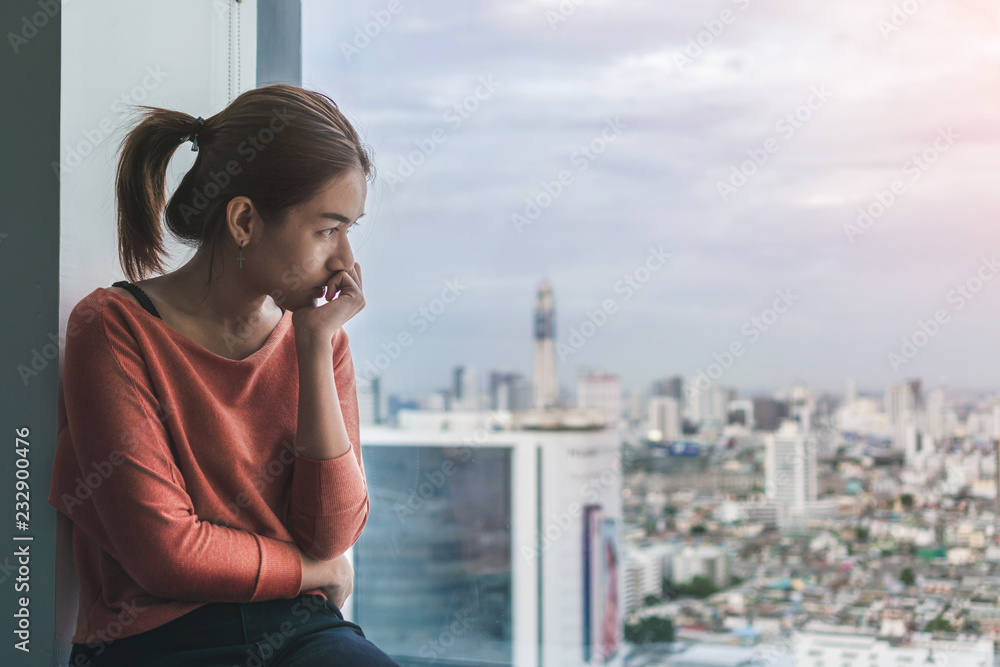 Fototapeta premium PTSD Mental health concept, Young depressed asian woman sitting alone near window in dark at evening time with low light environment.Selective focus.