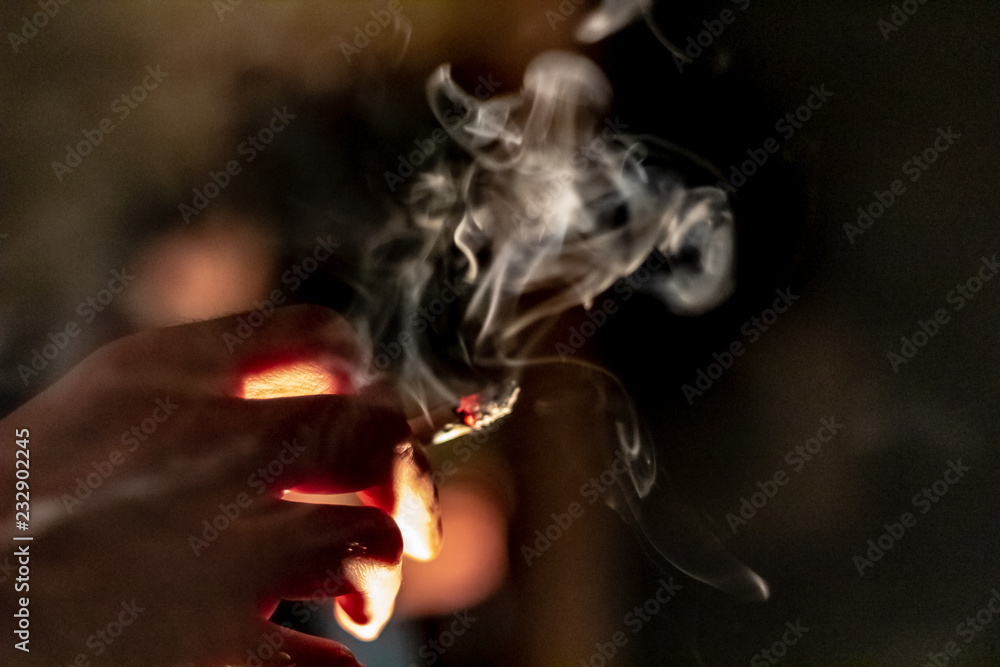 A hand holding a marijuana cigarette or joint with a large plume of thick smoke coming from it.