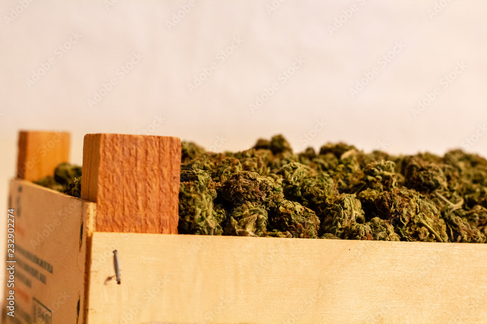 The corner of a wooden crate full of marijuana buds. The corner piece of wood making the top of the crate is visible with a large mound of marijuana buds next to it.