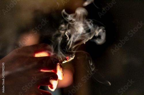 A hand holding a marijuana cigarette or joint with a large plume of thick smoke coming from it.