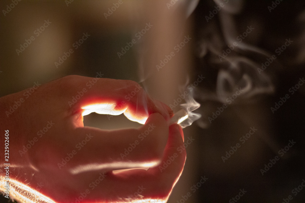 A hand holding a joint or marijuana cigarette while it burns sending smoke rising into the air around it.