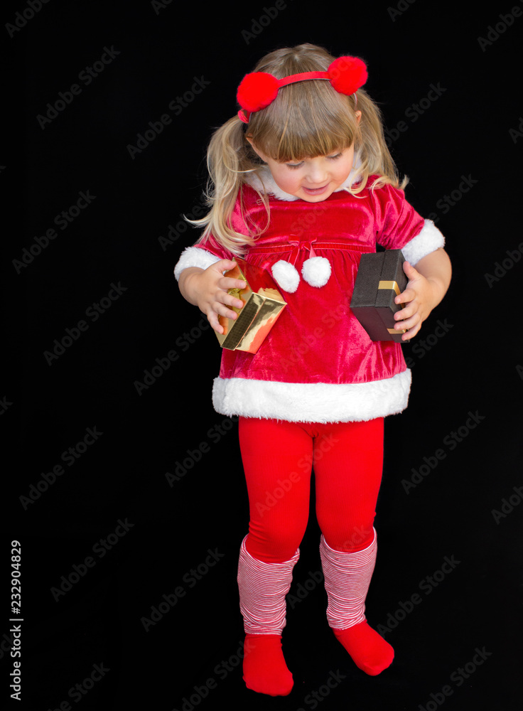 Little beautiful girl in a Christmas Santa suit, with fur balls on her head, holds gifts in her hands and rejoices. The concept of celebrating Christmas, New Year and gifts.