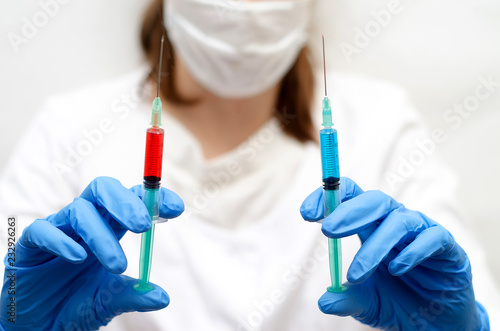 a woman in a white robe, mask and blue gloves, holding two syringes to choose from with blue and red liquid