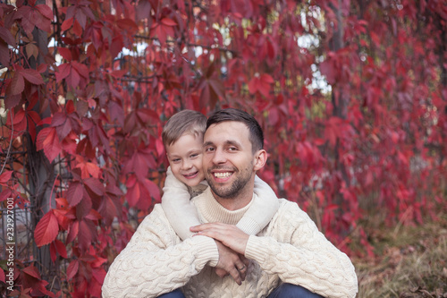 Dad and little son walk in the autumn park. Bright, warm autumn. Red leaves. Cozy. Portrait of dad and son in knitted sweaters.