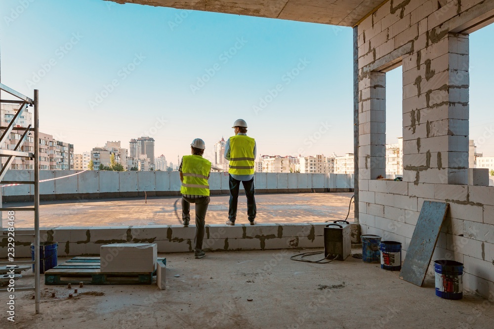 Two male builders at a construction site. Men looking into the distance to the panorama of the city. Building, development, teamwork and people concept Two male builders at a construction site. Men looking into the distance to the panorama of the city. Building, development, teamwork and people concept