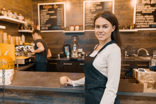 Portrait of young female small business owner - small modern coffee shop, confident woman standing at the counter with barista working in background making drinks