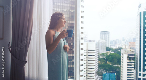 Beautiful young brunette woman drinking coffee by the panoramic window with amazing city view