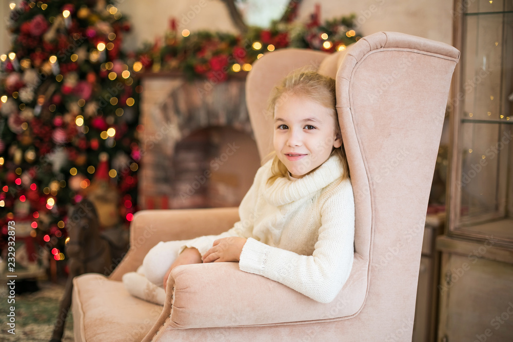 Christmas portrait of happy blonde child girl in white sweater siting on the floor near the Christmas tree. New Year Holidays Christmas portrait of happy blonde child girl in white sweater siting on the floor near the Christmas tree. New Year Holidays