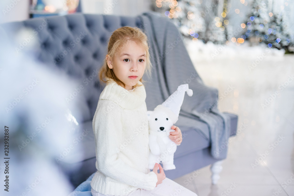 Merry Christmas and Happy New Year! Charming little blonde girl in white warm sweater with toy bear sitting on the blue armchair near Christmas tree Merry Christmas and Happy New Year! Charming little blonde girl in white warm sweater with toy bear sitting on the blue armchair near Christmas tree