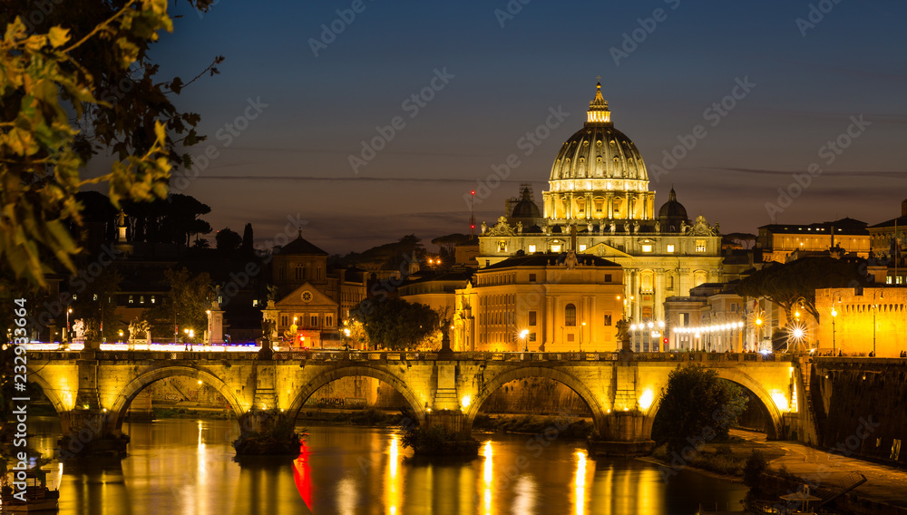 Illuminated St. Peter's Cathedral in Rome, Italy, and the Tiber River against dark sky at blue hour after sunset.
