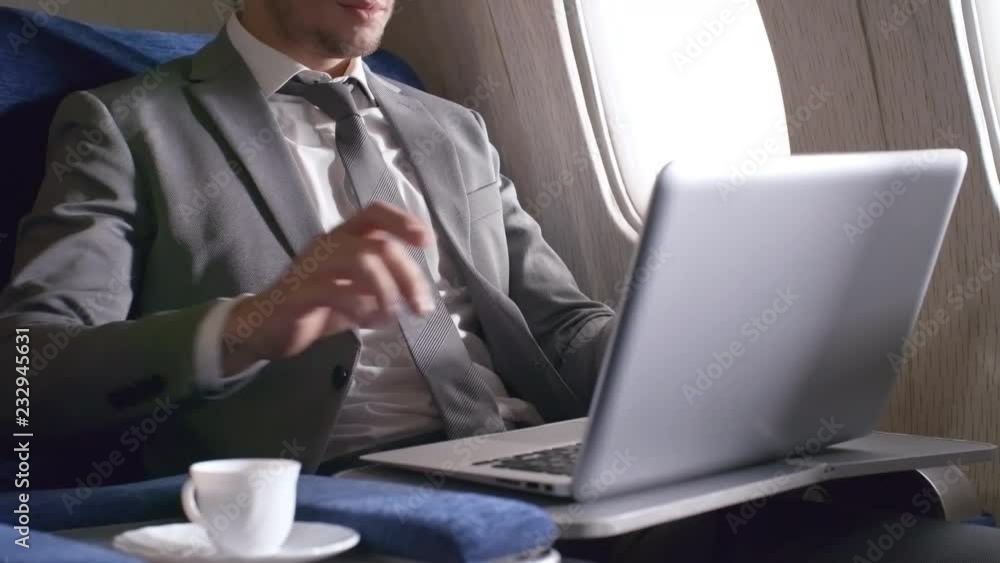 Tilt up shot of confident businessman in suit and necktie drinking coffee and working on laptop while traveling by plane Tilt up shot of confident businessman in suit and necktie drinking coffee and working on laptop while traveling by plane