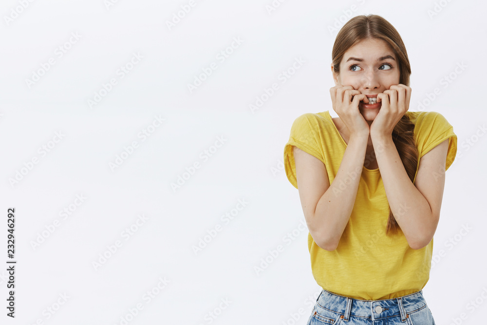 Waist-up shot of concerned nervous and anxious cute young timid woman in yellow t-shirt biting fingernails looking scared at upper left corner frightened, overreacting posing against white background Waist-up shot of concerned nervous and anxious cute young timid woman in yellow t-shirt biting fingernails looking scared at upper left corner frightened, overreacting posing against white background