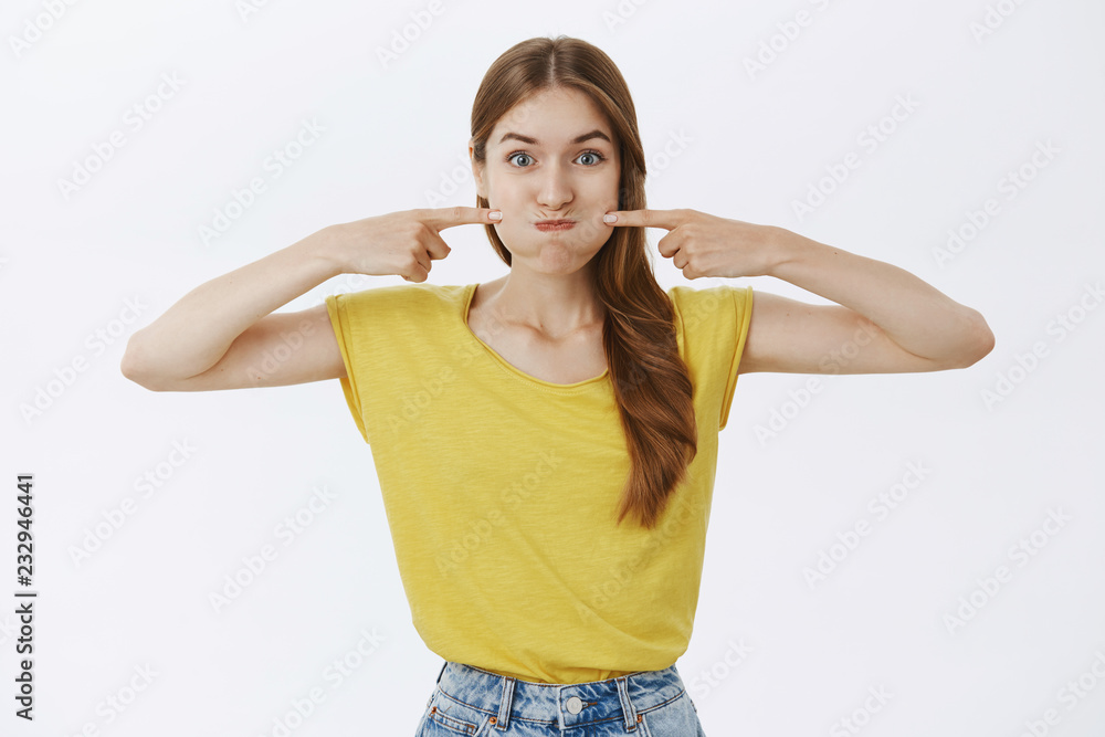 Fototapeta premium Charismatic funny and happy charming young female in yellow t-shirt pouting poking cheeks with index fingers and smiling as if holding breath playfully fooling around over white background