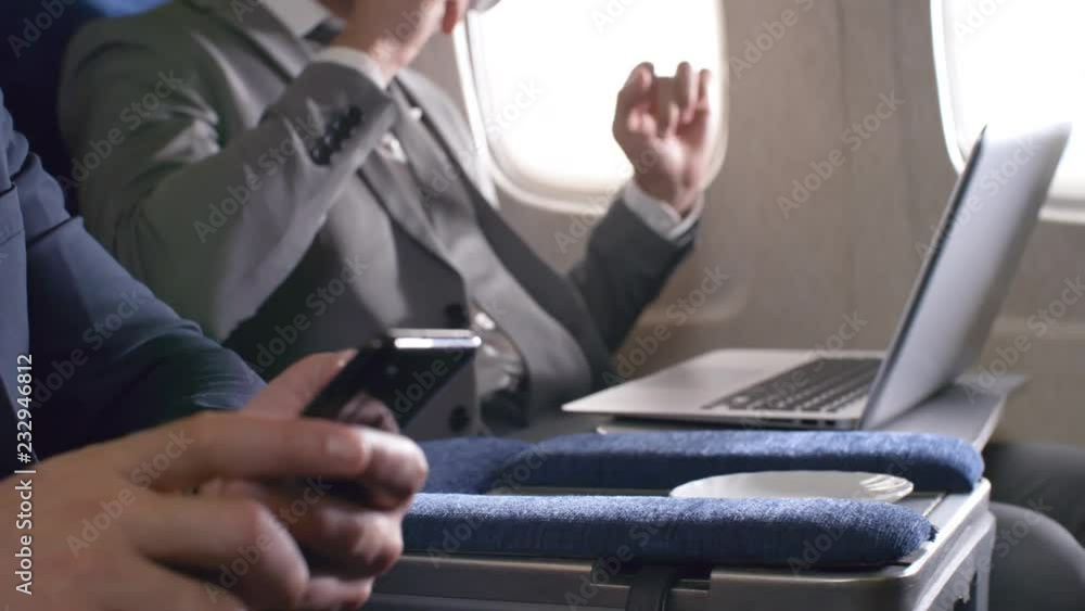 Mid-section shot of unrecognizable businesspeople in suits traveling by plane. Man in aisle seat typing on mobile phone as his busy colleague sitting by window working on laptop and drinking coffee Mid-section shot of unrecognizable businesspeople in suits traveling by plane. Man in aisle seat typing on mobile phone as his busy colleague sitting by window working on laptop and drinking coffee