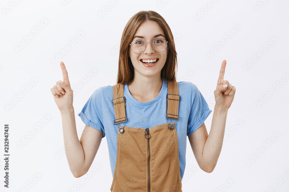 Charming friendly-looking charismatic female in glasses and trendy brown overalls pointing up with raised arms and smiling with joy and happiness posing over gray background delighted and amused Charming friendly-looking charismatic female in glasses and trendy brown overalls pointing up with raised arms and smiling with joy and happiness posing over gray background delighted and amused