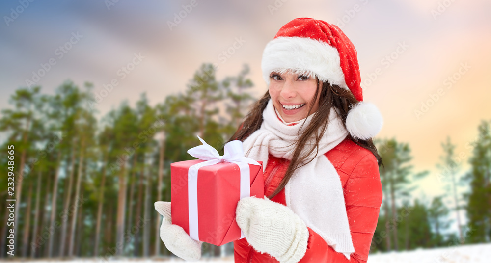 Obraz premium holidays and people concept - happy woman in santa hat with chrismas gift over winter forest background