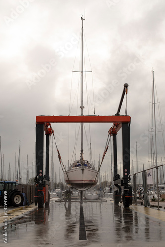 Fototapeta Naklejka Na Ścianę i Meble -  A boat lift is used to haul out a sailing boat for the winter at boat yards, the hull of the boat is cleaned with a high-pressure spray on a rainy cloudy day.