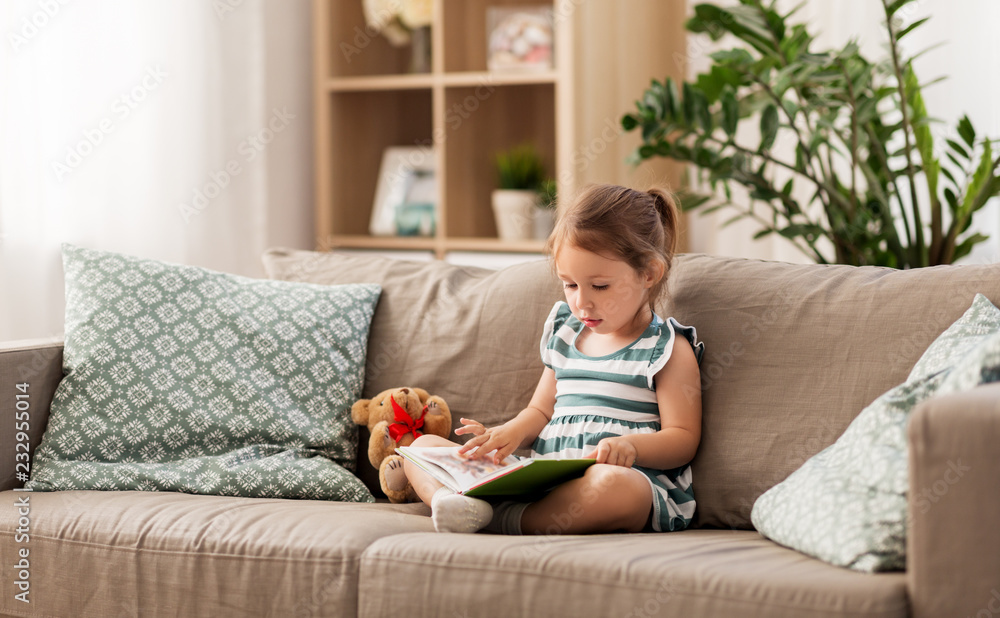childhood and people concept - little girl sitting on sofa reading book and toy teddy bear at home childhood and people concept - little girl sitting on sofa reading book and toy teddy bear at home