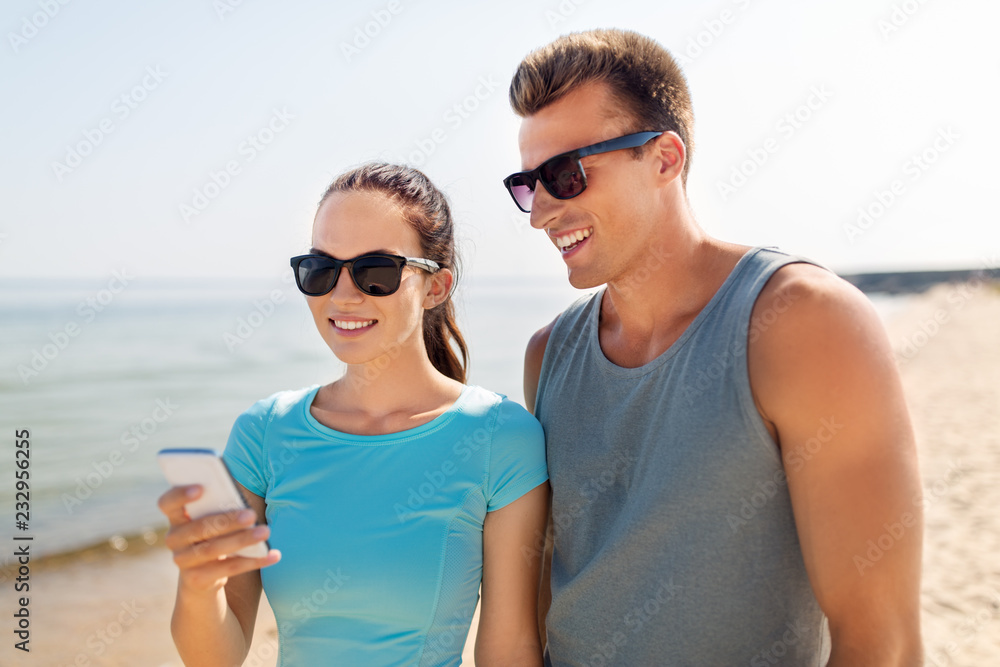 fitness, sport and lifestyle concept - happy couple in sports clothes and sunglasses with smartphones on beach