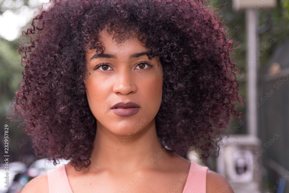 Portrait of young woman looking at camera. In the streets, natural lighting. She is black, on her early twenties, Afro style frizzy hair. Portrait of young woman looking at camera. In the streets, natural lighting. She is black, on her early twenties, Afro style frizzy hair.