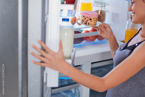 Woman breaking diet by taking cheesecake from fridge. Fridge full of groceries.