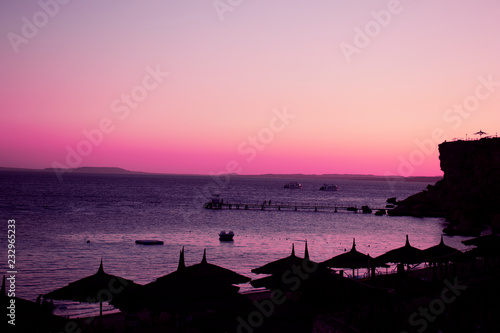 Beautiful evening view at the sea horizon. The silhouettes of parasols and beach are at the foreground. Amazing pink sky above dark sea.