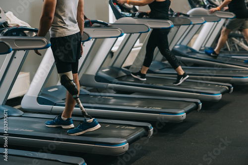cropped shot of man with artificial leg running on treadmills at gym with other people