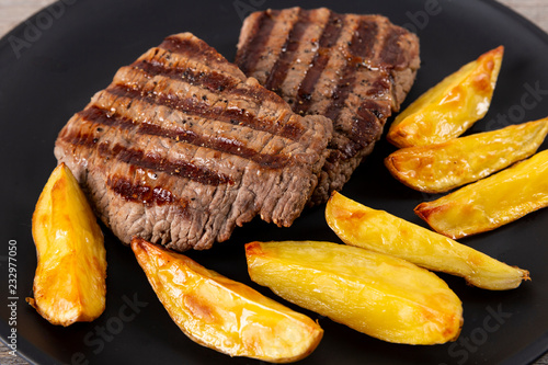 Two steaks and fried potatoes in a black plate on a wooden background.